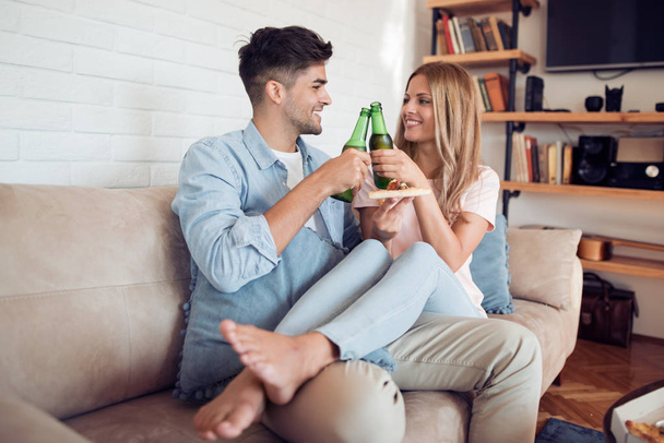 Young couple sitting on the couch in the living room, holding beer and smiling. - Fotó, kép