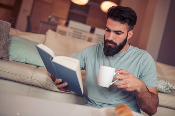 Jovem está sentado em um sofá e lendo um livro enquanto segura xícara de café
. - Foto, Imagem