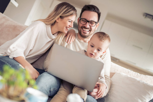 Gyönyörű fiatal család használ laptop, beszél, és mosolyogva, miközben ül a kanapén, otthon. - Fotó, kép