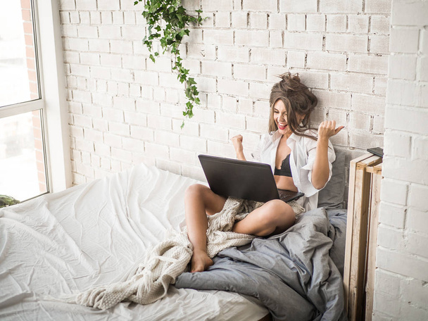 Ευτυχισμένη περιστασιακή όμορφη γυναίκα που εργάζεται σε ένα φορητό υπολογιστή κάθεται στο κρεβάτι στο σπίτι. - Φωτογραφία, εικόνα