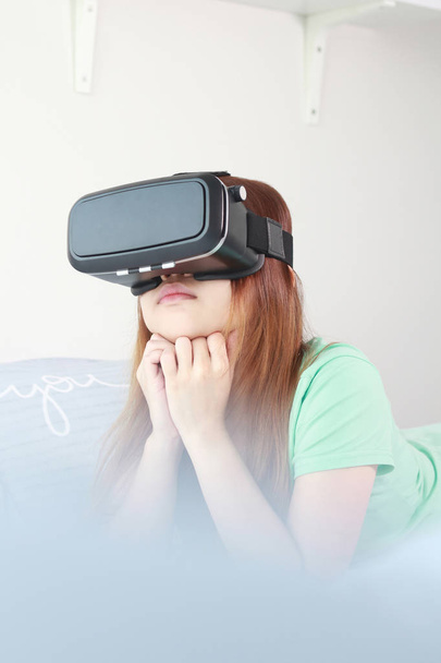 Νεαρή γυναίκα που φοράει γυαλιά εικονικής πραγματικότητας στο σπίτι. Ένα σετ κεφαλής εικονικής πραγματικότητας είναι ένα κεφάλι-mounted συσκευή που παρέχει εικονικής πραγματικότητας για τον κομιστή. - Φωτογραφία, εικόνα