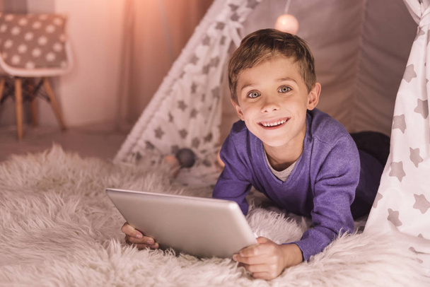 Digital tech. Cheerful happy nice boy lying in a tent and looking at you while holding a tablet - Fotografie, Obrázek