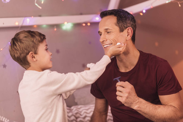 Positive cute boy putting foam on his fathers face - Photo, Image