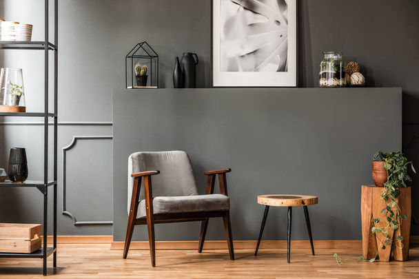 Grey armchair next to a wooden table in living room interior with plant and poster - Foto, imagen