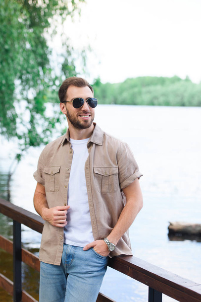 sonriente hombre guapo apoyado en la barandilla cerca del lago en el parque
 - Foto, imagen