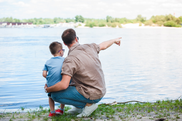 back view of father pointing on something at river to son at park - Fotoğraf, Görsel