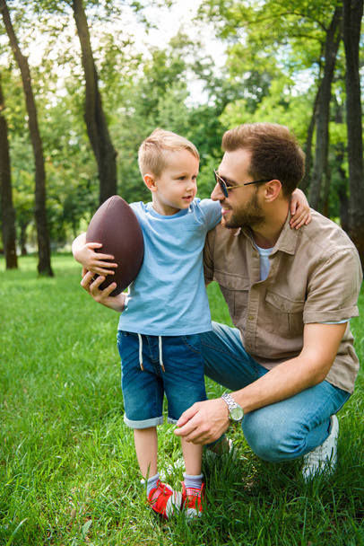 Ojciec i syn, przytulanie i trzymając piłkę futbolu amerykańskiego w parku - Zdjęcie, obraz