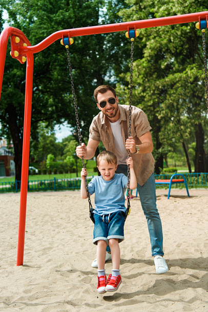 Isä ja poika hauskaa keinu leikkipaikka puistossa ja katsot kameraa
 - Valokuva, kuva