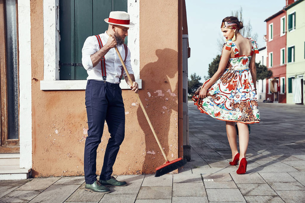 Beautiful sexy woman in colorful dress, headband and red heels and man with a broom, dressed in polka dot shirt, linen hat, bow tie and dark pants with suspenders, near colorful houses in Burano Island, Venice, Italy   - Fotó, kép