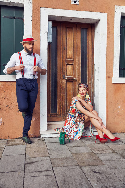 Fashionable couple near house in Burano Island, Venice, Italy. Woman in colorful dress and headband with big earring and green straw bag, red heels, sitting on stair, man in white shirt, linen hat, pants with suspenders stands next to her - Fotó, kép