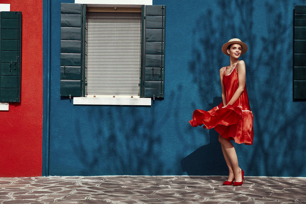 Mulher nova Fashoinable no vestido vermelho e saltos com chapéu de palha na cabeça, posando perto de casas azuis e vermelhas em Ilha de Burano. Com sombra de árvore na parede
 - Foto, Imagem