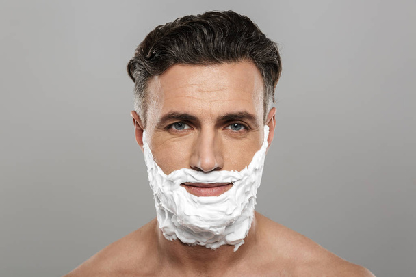 Photo of mature man standing isolated over grey wall background naked with shaving cream on face. - Foto, afbeelding