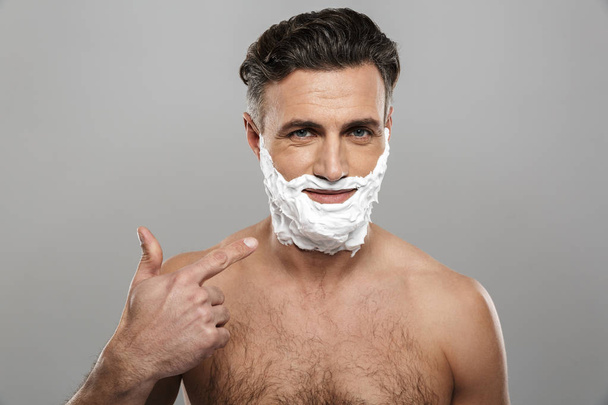 Image of mature man standing isolated over grey wall background naked with shaving cream on face pointing. - Φωτογραφία, εικόνα