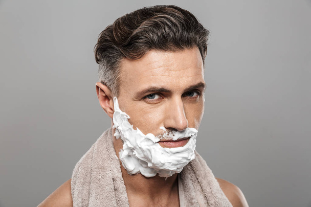 Photo of mature man standing isolated over grey wall background naked with shaving cream on face holding towel. - Foto, immagini