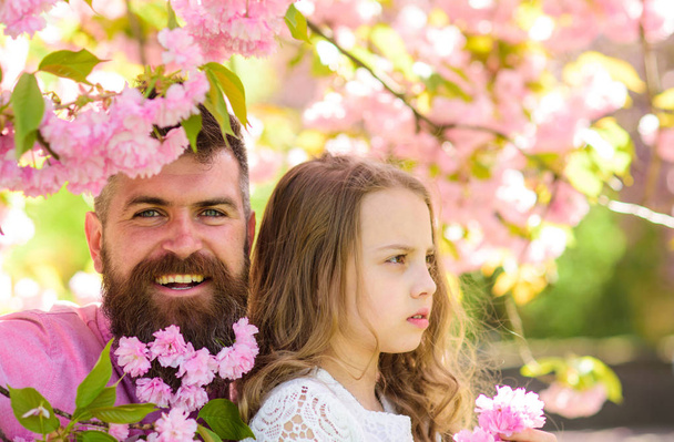 Fatherhood concept. Girl with dad near sakura flowers on spring day. Child and man with tender pink flowers in beard. Father and daughter on happy and calm faces , sakura background - Foto, imagen