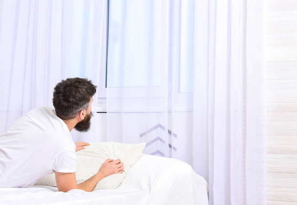 Man in shirt laying on bed awake, white curtain on background. Wake up and morning concept. Macho with beard and mustache relaxing after nap. Guy waking up in morning, looking at windo. - Photo, image