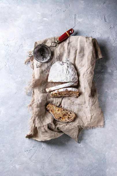 Sliced Christmas cake, traditional German festive baking. Wholegrain stollen with raisins and sugar powder on linen napkin with vintage sieve over gray texture background. Top view, space - Foto, Bild