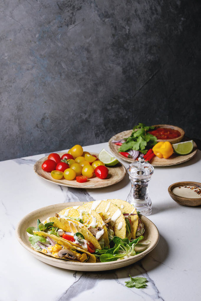 Variety of vegetarian corn tacos with vegetables, green salad, chili pepper served on ceramic plate with tomato and cream sauces with ingredients above on white marble kitchen table - Foto, Bild
