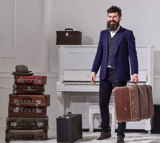 Macho elegant on smiling face stands near pile of vintage suitcase, holds suitcase. Man, traveller with beard and mustache with luggage, luxury white interior background. Baggage delivery concept - Φωτογραφία, εικόνα