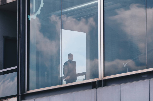 άνθρωπος των επιχειρήσεων που αναζητούν πάνω από το παράθυρο σε ένα σύγχρονο κτήριο με αντανάκλαση του ουρανού και τα σύννεφα στα παράθυρα - Φωτογραφία, εικόνα