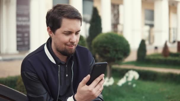 Adult man is using video calling by smartphone, sitting outdoors - Séquence, vidéo