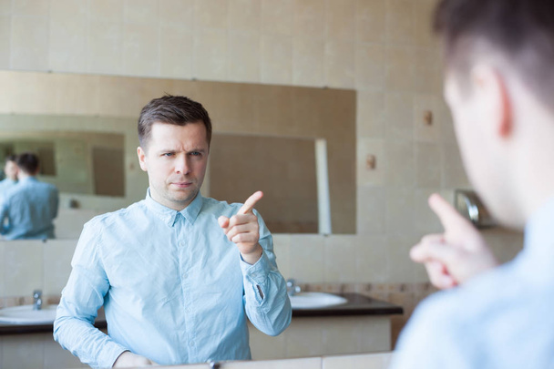 Um jovem de camisa azul ajusta o cabelo em frente ao espelho na sanita. Preocupa-se e prepara-se para o encontro
 - Foto, Imagem