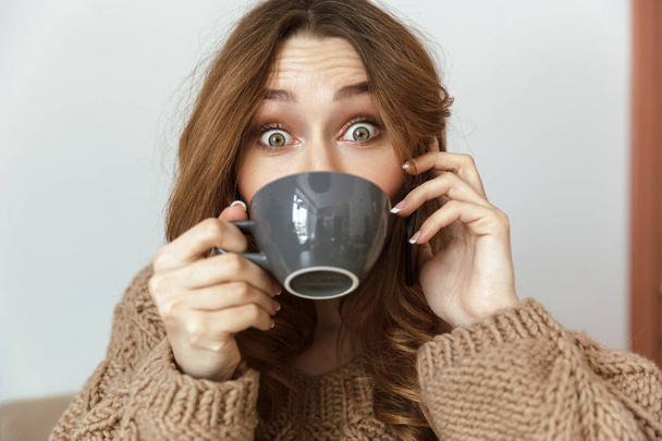 Photo of cheerful young woman 20s looking on camera with exciting look and drinking cup of tea at home or in cafe - Foto, afbeelding