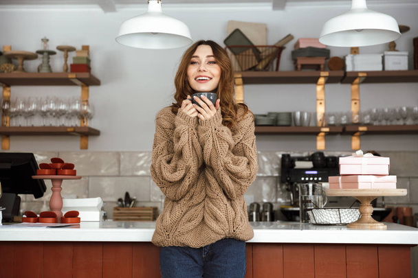 Photo of happy woman in sweater standing near buffet in cafe and smiling while holding cup of tea or coffee - Fotoğraf, Görsel