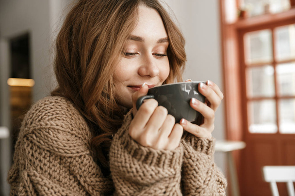 Portrait closeup of glad smiling woman in knitted sweater sitting at table in cafe and drinking cup of tea or coffee - Foto, Imagem