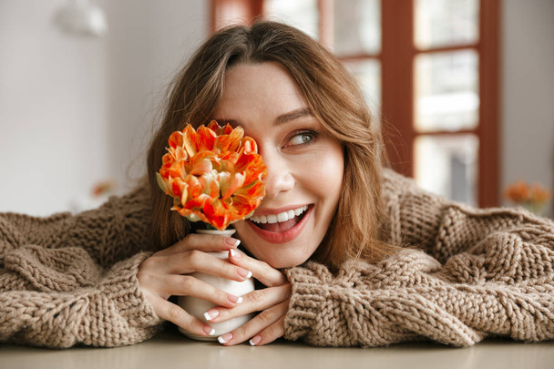 Beautiful photo of lovely woman in sweater sitting at table in cafe looking aside with smile while holding flower close to face - 写真・画像