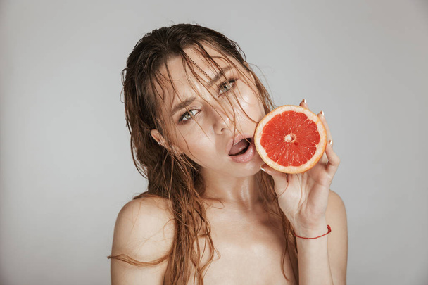 Fashion portrait of a topless beautiful woman with makeup and wet hair holding sliced grapefruit isolated over gray background - Zdjęcie, obraz