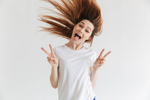 Happy screaming woman in t-shirt showing peace gestures and looking at the camera over grey background - Fotó, kép