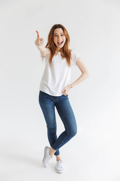 Full length image of Happy woman in t-shirt posing with arm on hip while showing thumb up and looking at the camera over grey background - Fotó, kép