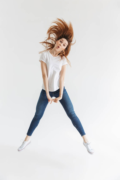 Full length image of Playful woman in t-shirt jumping and looking at the camera over grey background - Foto, imagen