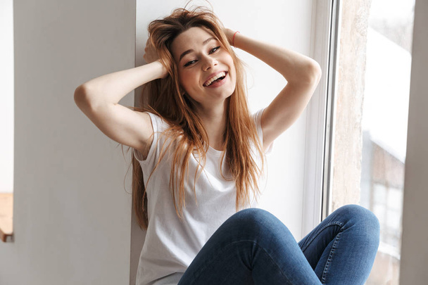 Cheerful woman in t-shirt sitting on windowsill while holding her head and looking at the camera over grey background - Foto, Bild