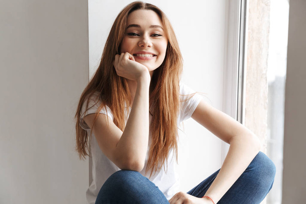 Smiling woman in t-shirt sitting on windowsill while looking at the camera at home - Foto, Imagen