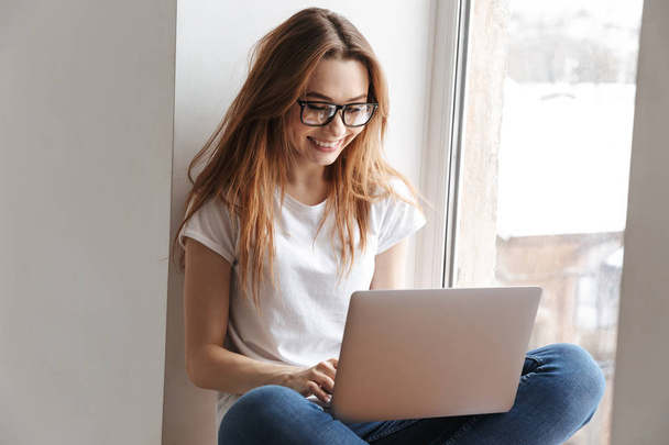Smiling woman in t-shirt and eyeglasses sitting on windowsill while using laptop coputer - Fotó, kép