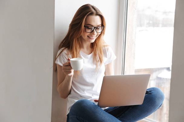 Pleased woman in t-shirt and eyeglasses sitting windowsill while using laptop computer and holding cup of coffee - Foto, Bild