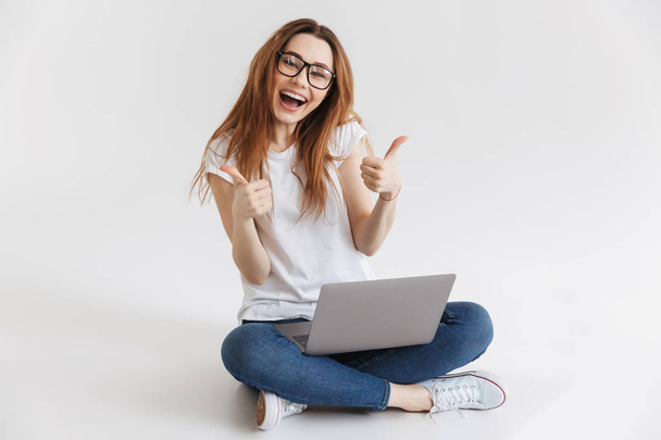 Happy woman in t-shirt and eyeglasses sitting on the floor with laptop computer while showing thumbs up and looking at the camera - Zdjęcie, obraz