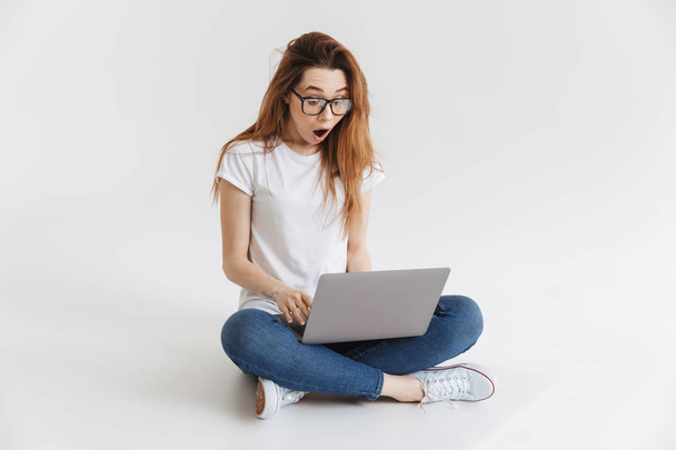 Shocked woman in t-shirt and eyeglasses sitting on the floor and using laptop computer over grey background - Foto, Bild