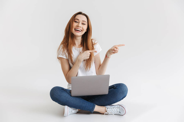glückliche Frau im T-Shirt, die mit Laptop auf dem Boden sitzt, wegzeigt und vor grauem Hintergrund in die Kamera schaut - Foto, Bild