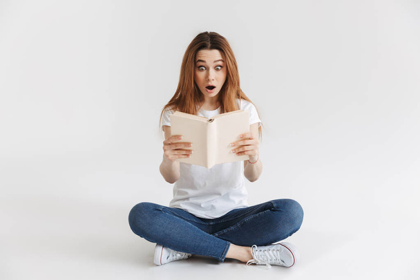Surprised woman in t-shirt sitting on the floor and reading book with open mouth over grey background - Photo, Image
