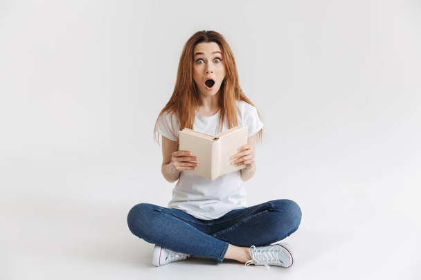Shocked woman in t-shirt sitting on the floor while holding book and looking at the camera over grey background - Photo, Image