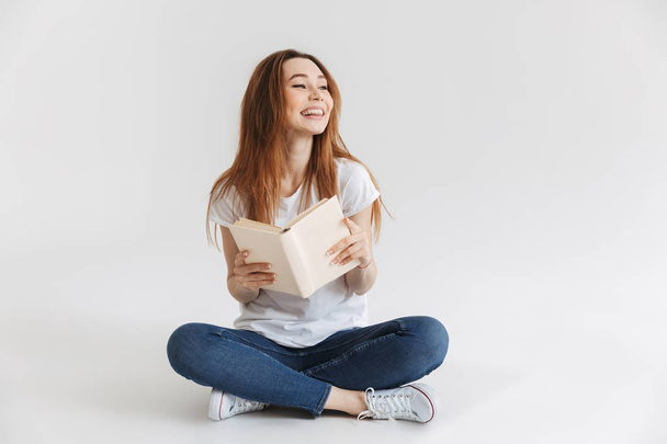 Portrait of a smiling young girl holding book while sitting with legs crossed isolated over white background - Foto, Imagen