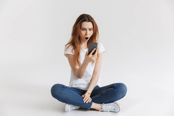 Portrait of a shocked young girl looking at mobile phone while sitting with legs crossed isolated over white background - 写真・画像