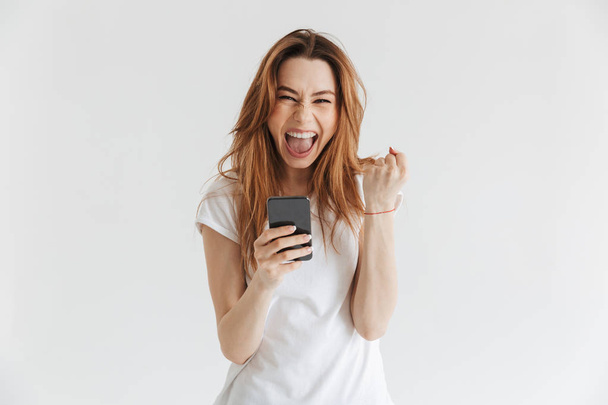 Happy woman in casual clothes holding smartphone while rejoices and looking at the camera over grey background - Foto, Imagen