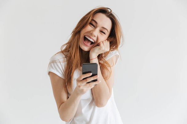 Cheerful woman in casual clothes holding smartphone and looking at the camera over grey background - Fotó, kép