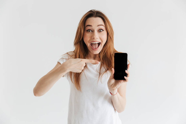 Surprised happy woman in casual clothes showing blank smartphone screen and pointing on it while looking at the camera over grey background - Foto, Imagen
