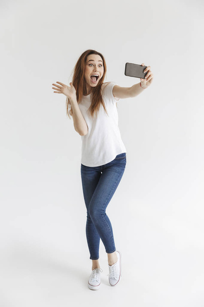 Full length image of Joyful woman in casual clothes posing and making selfie on smartphone while waving at camera over grey background - Фото, зображення