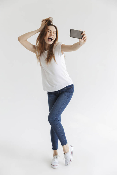 Full length image of Joyful woman in casual clothes posing and making selfie on smartphone over grey background - Foto, Bild